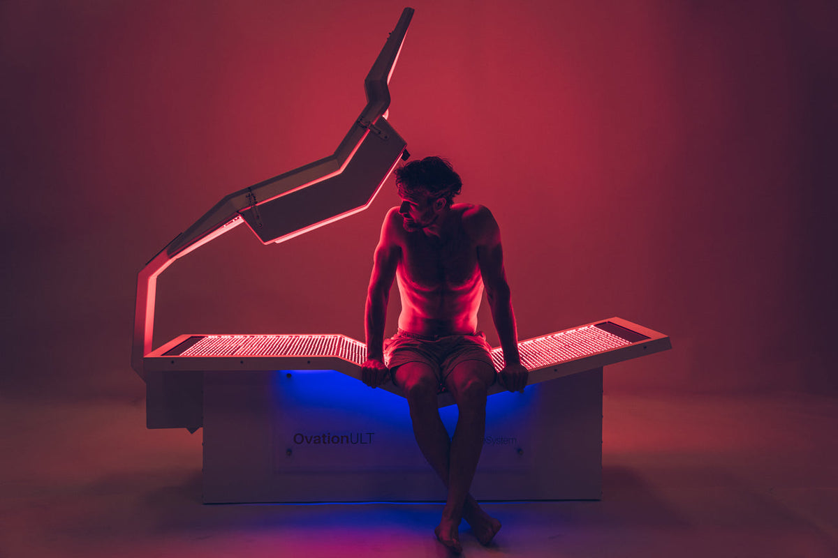 Man sitting on the OvationULT red light therapy bed in a glowing red and blue lit room.