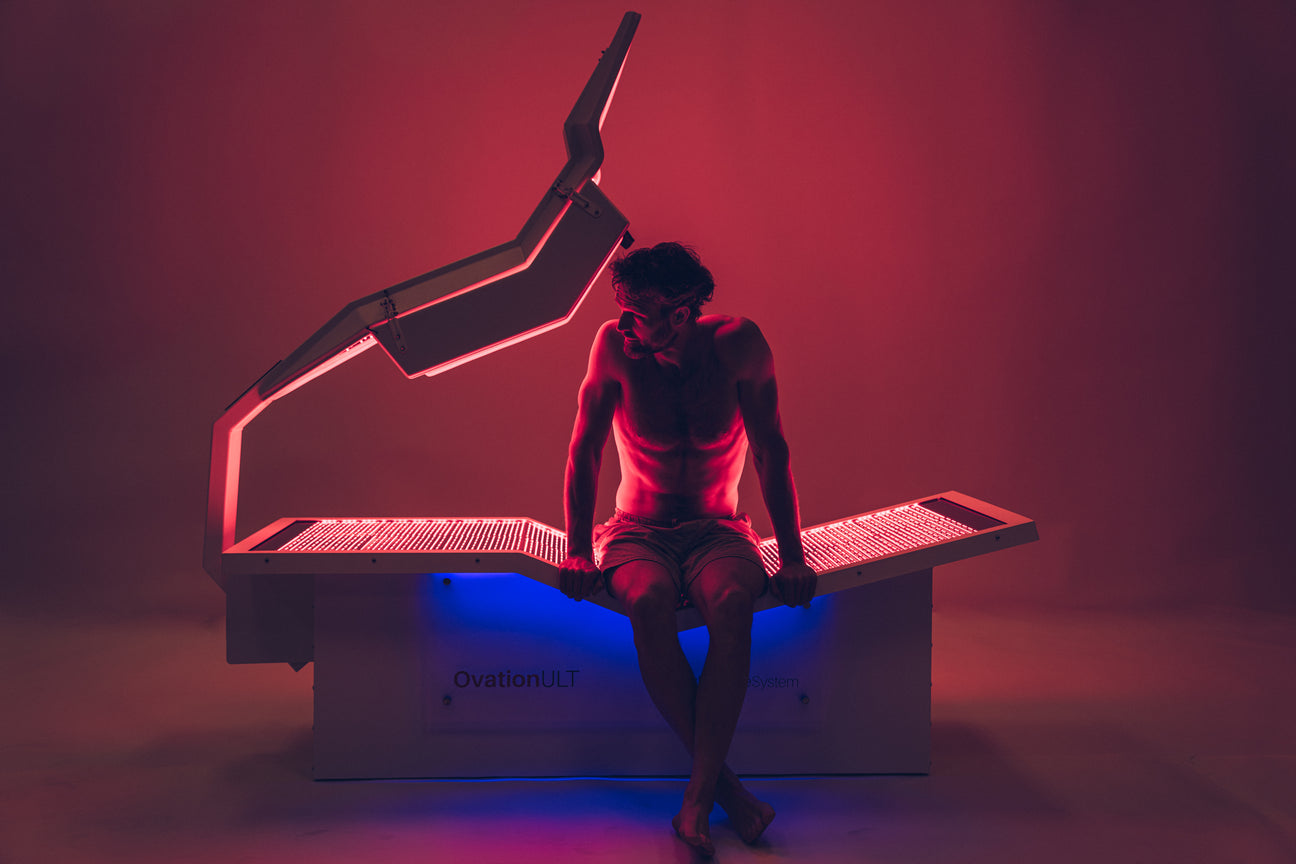 Man sitting on the OvationULT red light therapy bed in a glowing red and blue lit room.